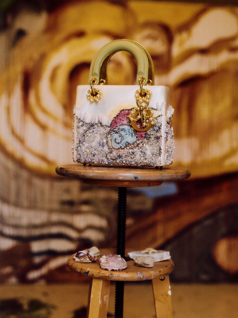 Close-up of a Lady Dior handbag with embellished details, placed on a wooden stool, and set against an abstract painted backdrop.
