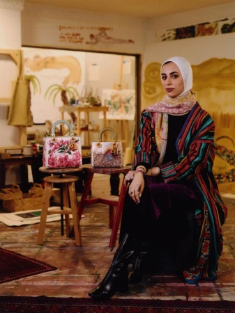 Woman in headscarf and colorful jacket poses in artist's studio near painted handbags and artwork with natural light, and red rug.