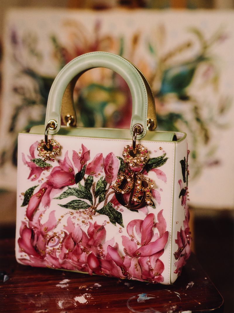 Elegant white handbag featuring a pink floral design with a pale green handle, gold accents, and hanging charm on a dark wood surface.