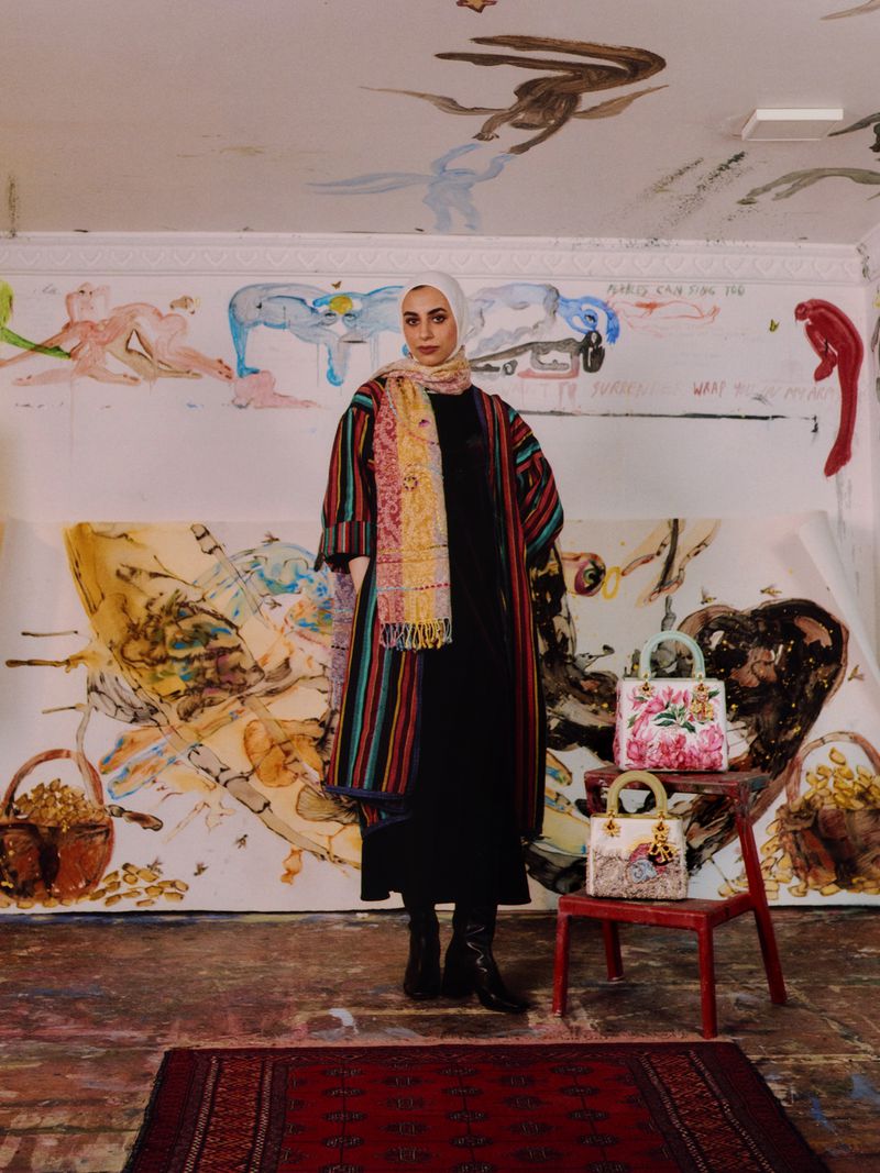 Young woman in hijab and striped coat poses in an art studio, next to floral purses on a stool, and an abstract painted backdrop and floor.