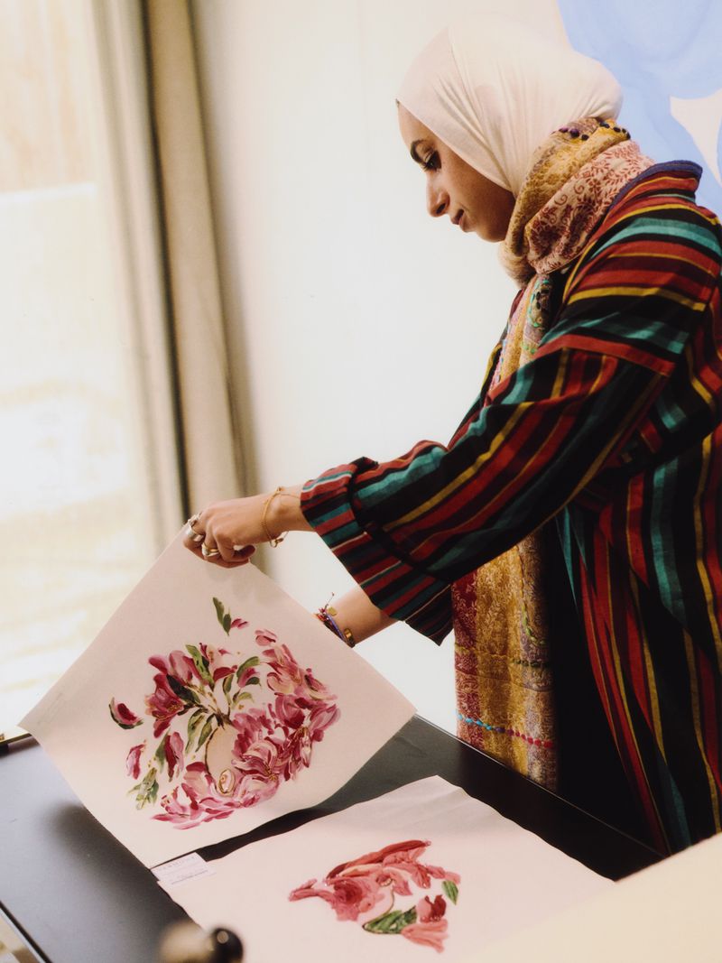 Adult female artist wearing a hijab creating watercolor painting of a flower bouquet in an indoor studio setting with natural light.
