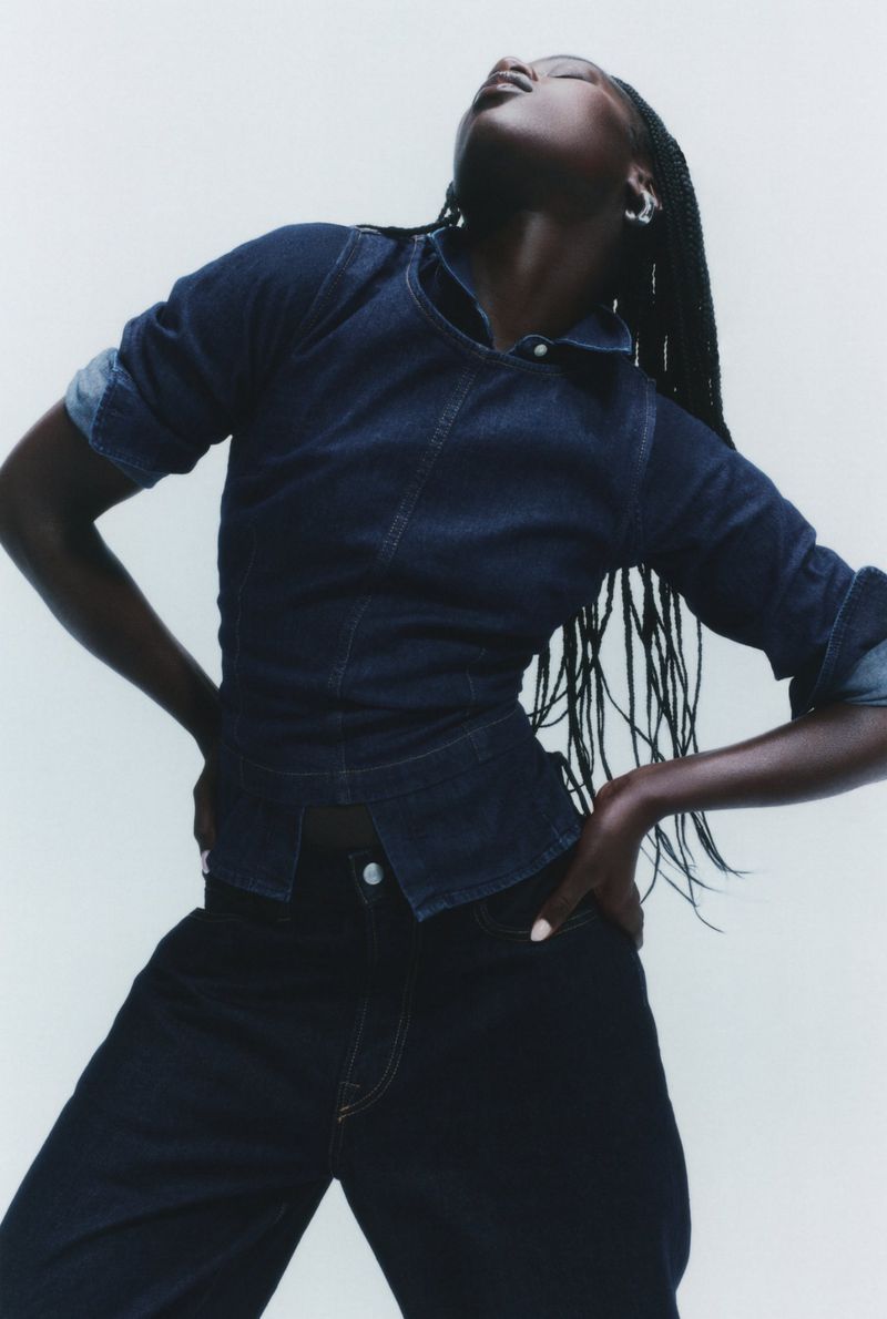 A dark-skinned adult woman is posing in a fitted denim shirt and dark jeans against a white background in a studio fashion shot.