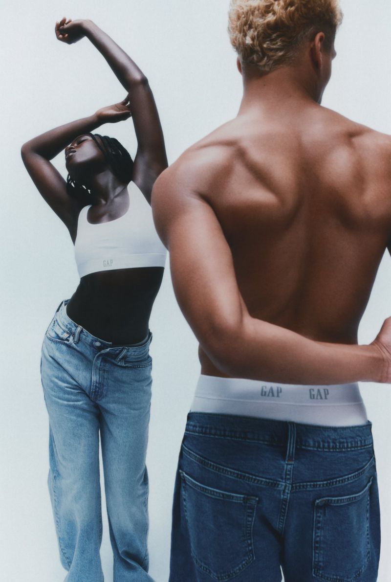 Two models wear Gap branded clothing in a bright studio shot. A woman in a white sports bra and jeans poses near a shirtless man.