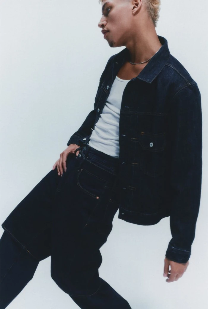 Model wearing a dark wash denim jacket and white tank top leaning back against a plain white studio backdrop.