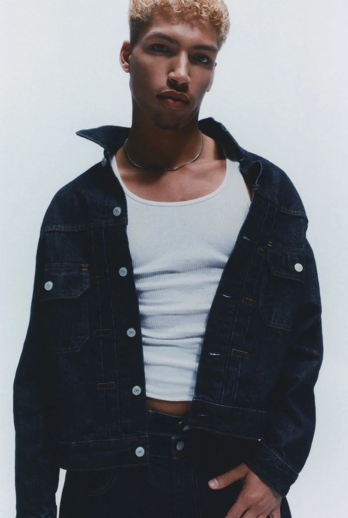 Young man wearing an open dark denim jacket over a white ribbed tank top posing against a clean white studio backdrop.