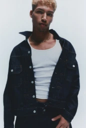 Young man wearing an open dark denim jacket over a white ribbed tank top posing against a clean white studio backdrop.