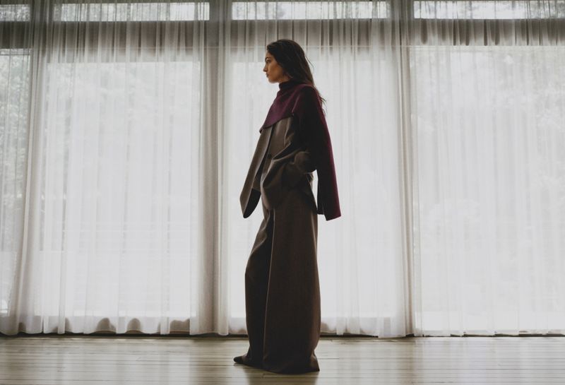 Woman in burgundy top and brown palazzo pants poses against a window with white curtains in a studio setting, looking left.