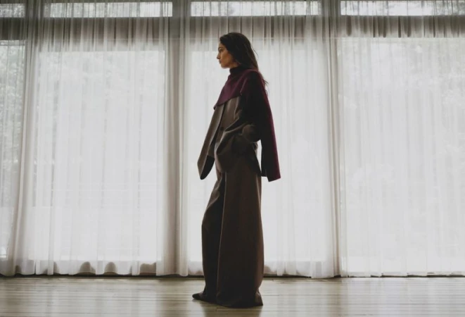 Woman in burgundy top and brown palazzo pants poses against a window with white curtains in a studio setting, looking left.