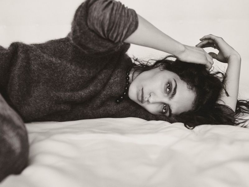 Monochrome portrait of a young woman lying on a white surface wearing a knit sweater and pearl necklace, looking at the camera.