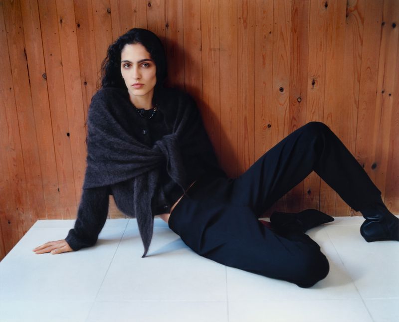 A dark-haired woman is lying down, wearing a dark sweater and black pants, posing in front of a paneled wooden wall.