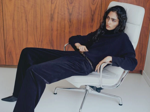 Adult female model in a relaxed pose in a white chair wearing black sweater and pants in an indoor studio with wood panel background.