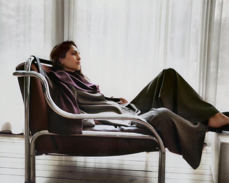 A woman reclines in a metal and leather lounge chair in a casual pose, wearing draped clothing in a naturally lit interior.
