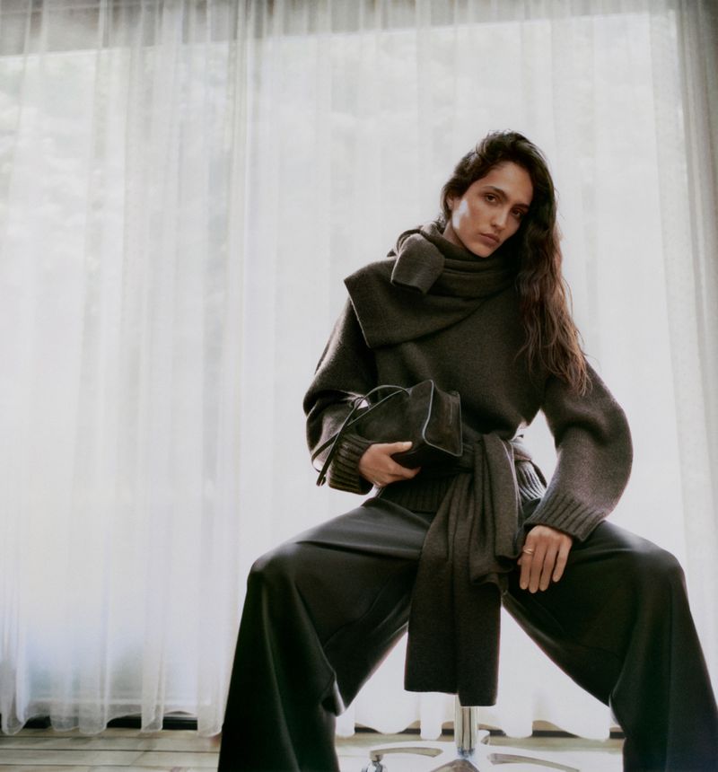 Elegant woman poses indoors wearing a dark grey sweater, scarf, and wide-leg trousers, holding a black handbag on her lap.