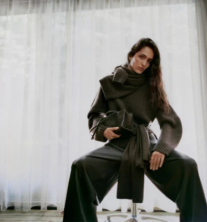 Elegant woman poses indoors wearing a dark grey sweater, scarf, and wide-leg trousers, holding a black handbag on her lap.