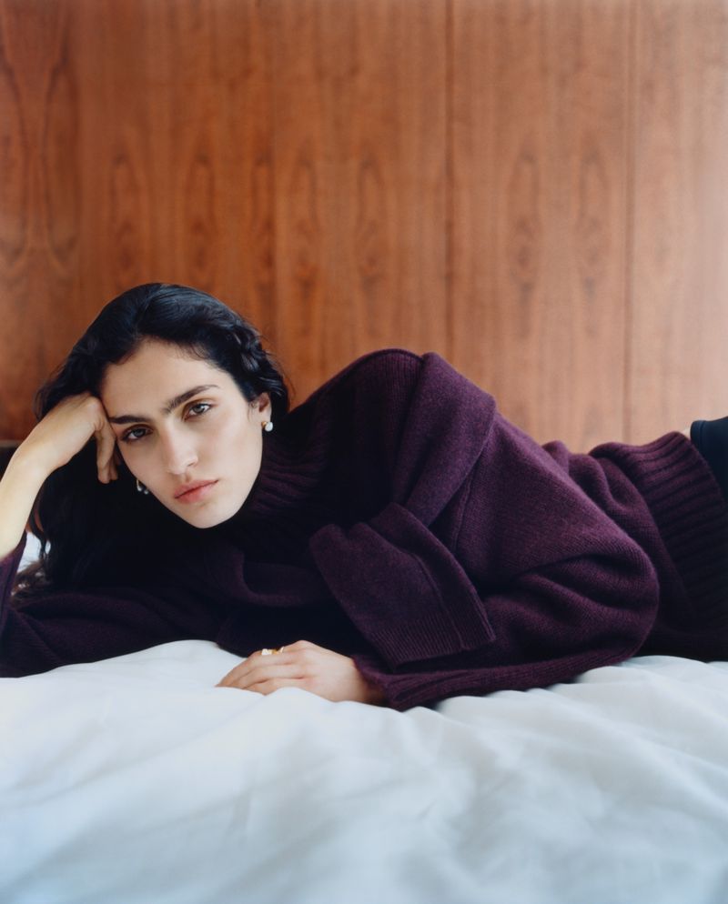 Sara Caballero lying on a white bed in a dark purple knit sweater in front of a wood-paneled wall.