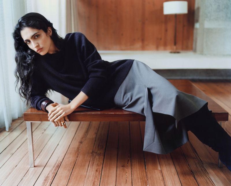 A woman with long dark hair lies across a wooden bench in a dark sweater and grey skirt in a softly lit interior fashion shot.
