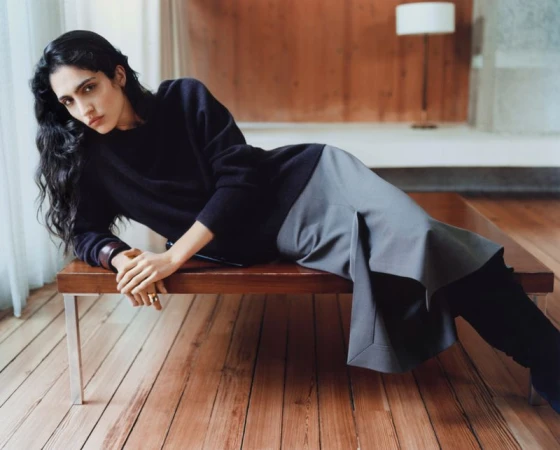 A woman with long dark hair lies across a wooden bench in a dark sweater and grey skirt in a softly lit interior fashion shot.
