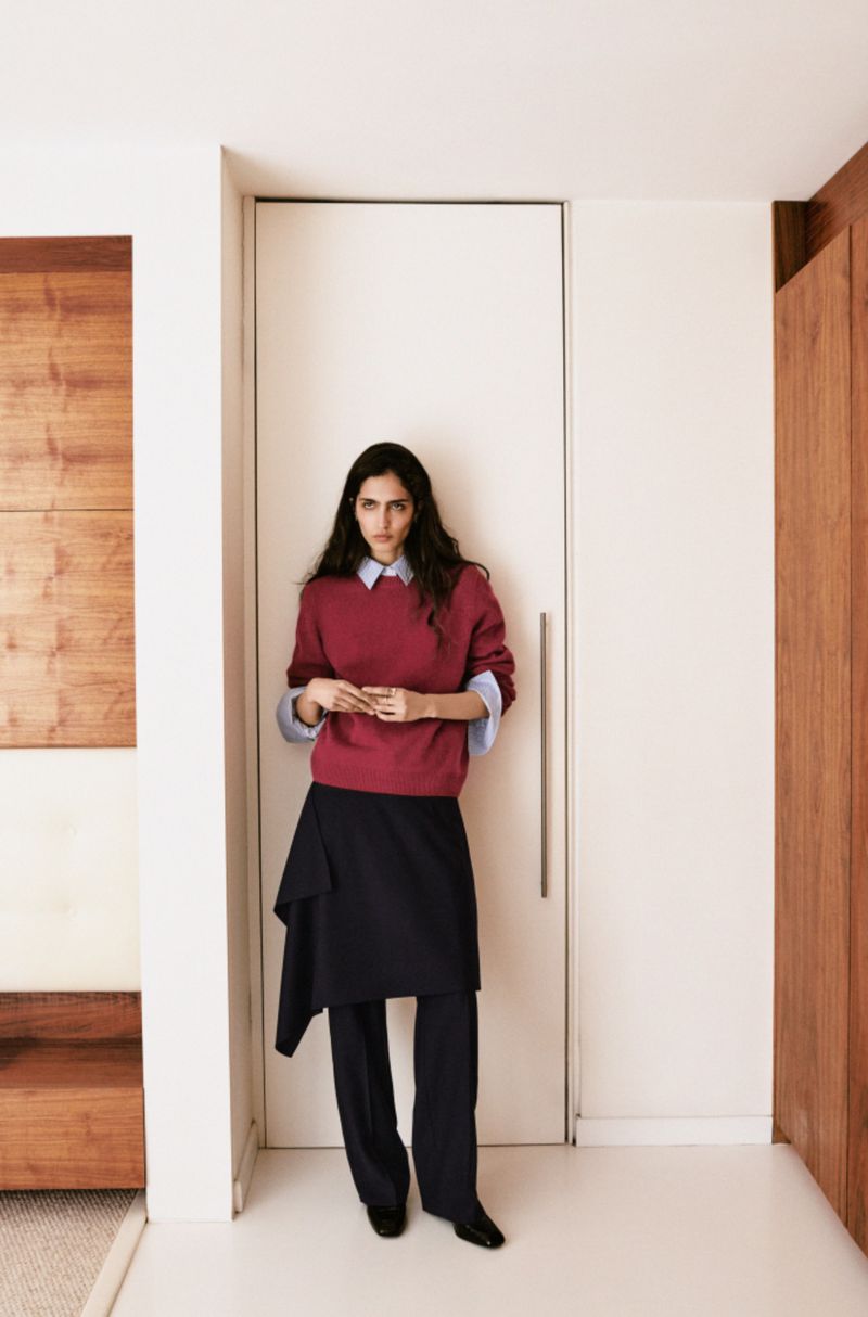 Indoor fashion shot of an adult female wearing a burgundy sweater, collared shirt, dark asymmetrical skirt, and dark pants.
