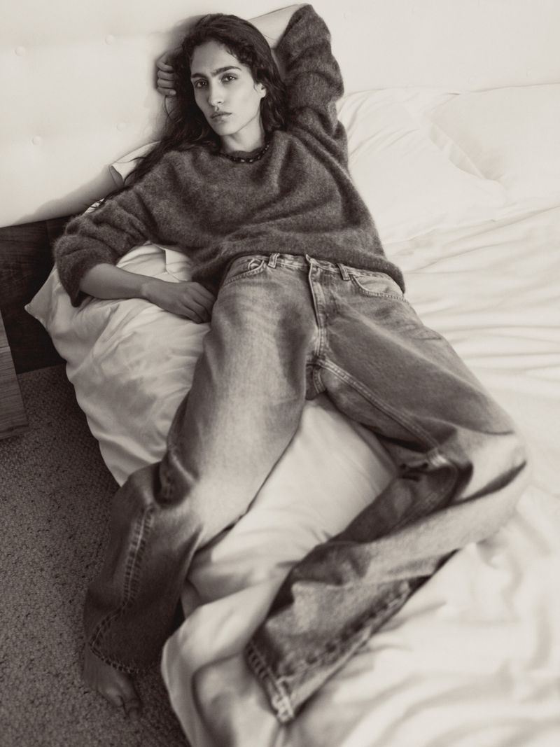Model Sara Caballero reclining on a bed wearing a fuzzy grey knit sweater and relaxed denim jeans in black and white.