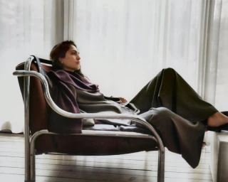Model relaxing in a leather chair wearing a grey coat and purple scarf near sheer curtains