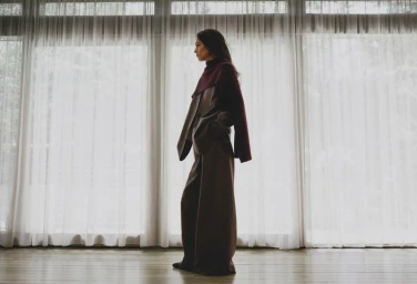 Model Sara Caballero stands in profile wearing wide-leg brown trousers and a burgundy knit layer before floor-to-ceiling sheer curtains.