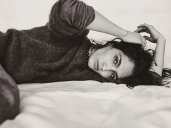 Model lying down on white sheets wearing a dark textured cashmere sweater with hands resting near her hair.