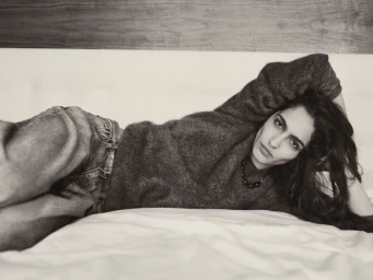 Model lying on a white bed wearing a dark cashmere sweater and jeans in a monochrome interior shot for COS Cashmere.