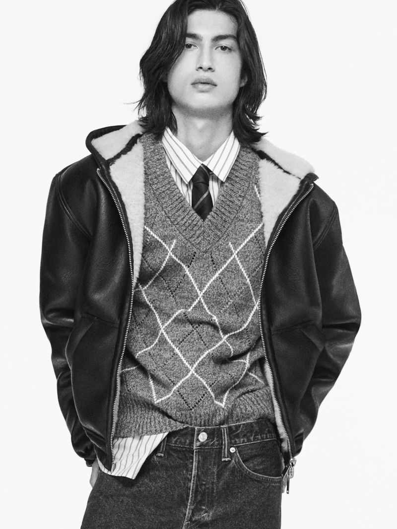 Studio portrait of a male fashion model. He is wearing a black leather jacket, grey sweater vest, striped shirt, and dark denim jeans.