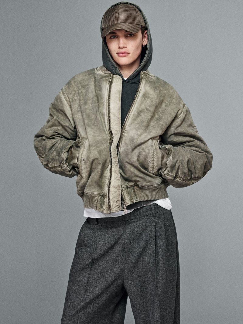 Young adult in hooded sweatshirt and green bomber jacket poses against a neutral gray backdrop in a fashion editorial shot.