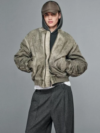 Young adult in hooded sweatshirt and green bomber jacket poses against a neutral gray backdrop in a fashion editorial shot.