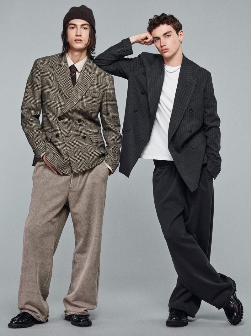 Two young adult men pose in a photo studio wearing business-casual attire, including blazers and loose-fitting pants, against a neutral grey background.