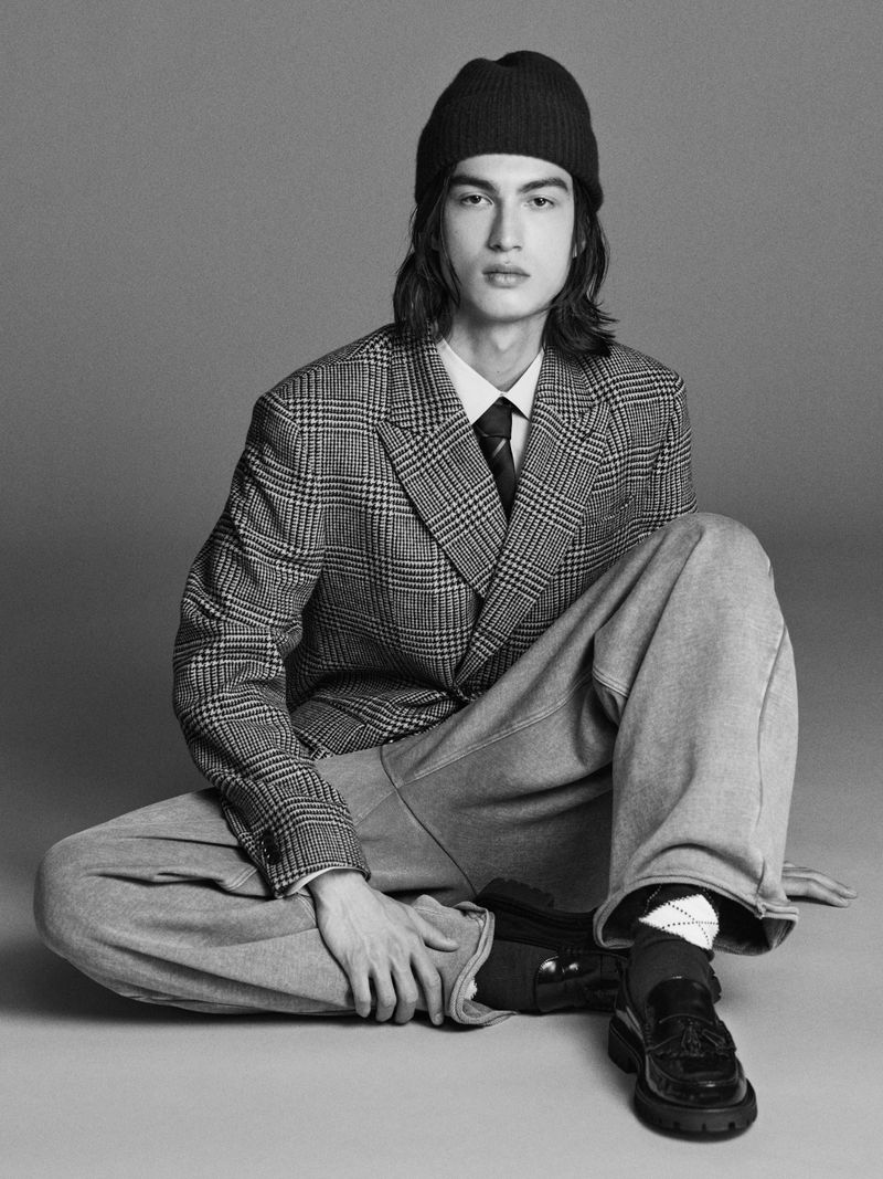 Man in plaid blazer, beanie, and wide-leg pants sitting on the floor in a fashion portrait with neutral grey background. Monochrome.