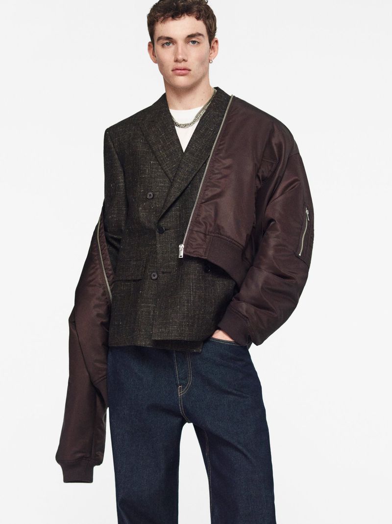 Fashion portrait of a slender adult male wearing a unique grey blazer with dark blue jeans against a white background in studio lighting.