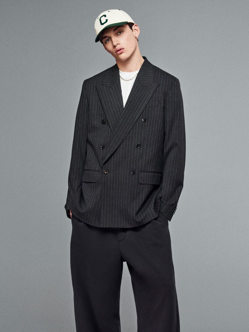 Portrait of a young adult male wearing a pinstripe blazer, dark trousers, baseball cap, and silver necklace against a grey backdrop.
