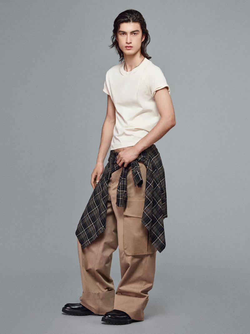 Full-shot of a young adult male posing in a studio with a casual outfit, tan pants, black shoes, and a plaid shirt tied around his waist.