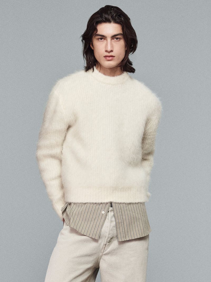 Studio shot of a young adult male with long dark hair wearing a fuzzy white sweater and light pants in front of a grey backdrop.