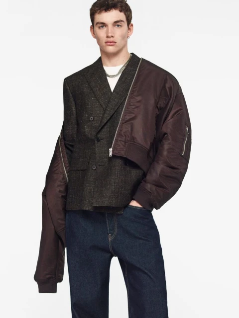 Male model in a textured tweed blazer, draped brown bomber jacket, and dark blue jeans against a white seamless background.