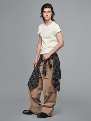 Male model wearing a white t-shirt, wide-leg beige cargo pants, and a plaid shirt tied around the waist against a grey background.