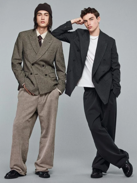Two male models posing in tailored blazers, wide-leg trousers, and chunky shoes against a plain grey studio backdrop.