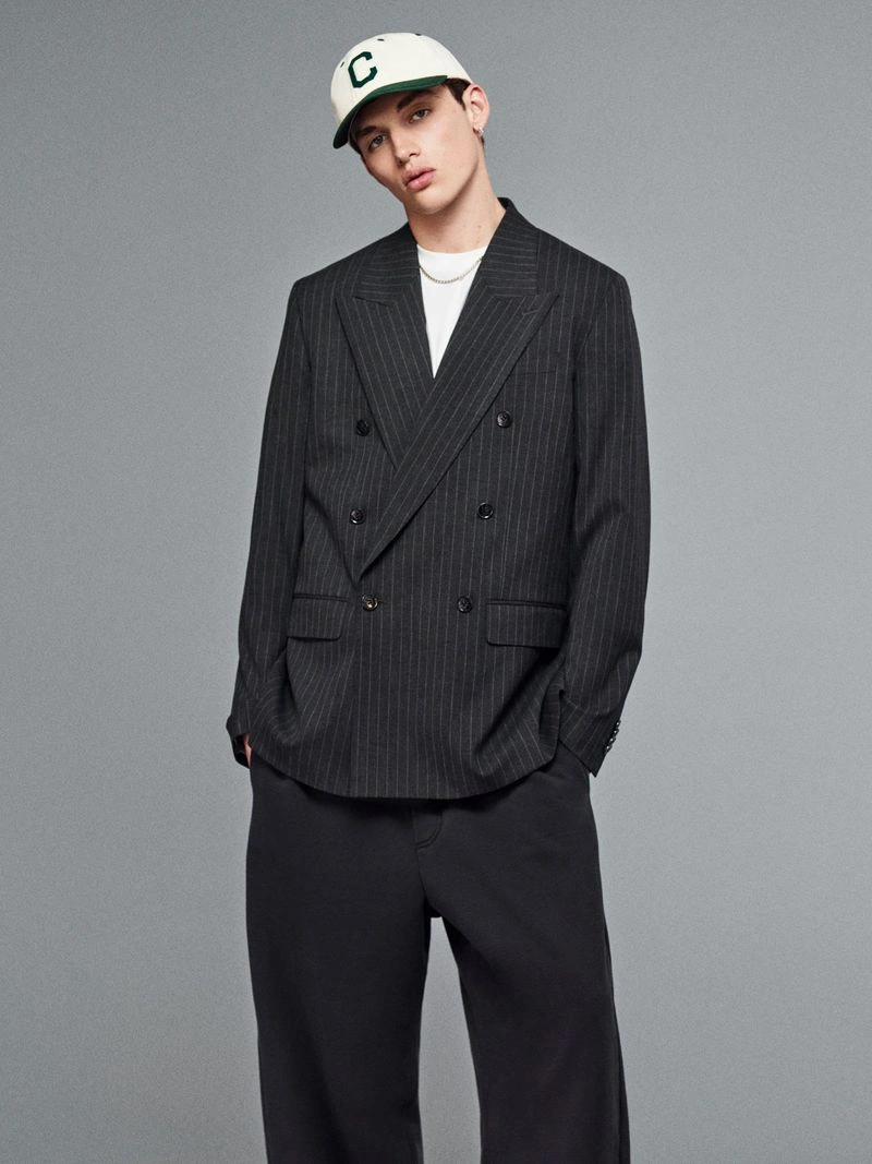 Male model wearing a dark pinstripe double-breasted blazer and a white baseball cap against a seamless grey backdrop.