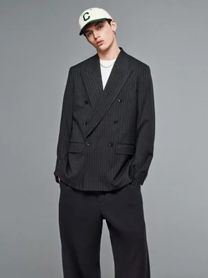 Male model wearing a dark pinstripe double-breasted blazer and a white baseball cap against a seamless grey backdrop.