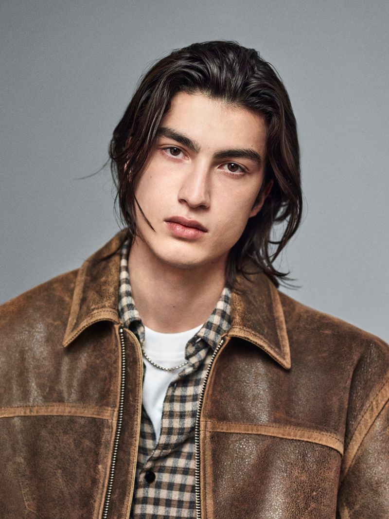 A young man with long dark hair wearing a distressed brown leather jacket and plaid shirt against a grey studio backdrop.
