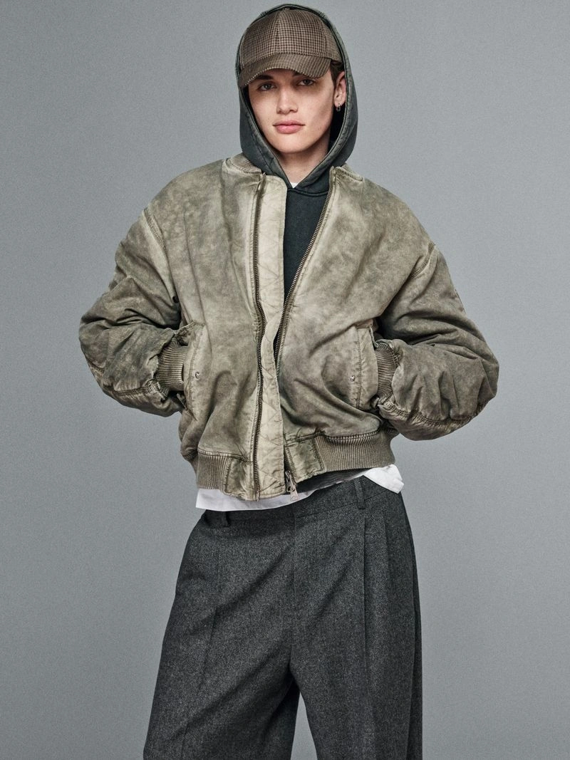 Model wearing a washed olive bomber jacket, grey hoodie, houndstooth baseball cap, and wide-leg pleated trousers against a grey backdrop.