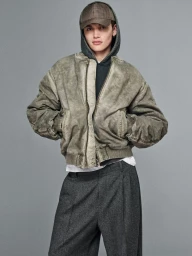 Model wearing a washed olive bomber jacket, grey hoodie, houndstooth baseball cap, and wide-leg pleated trousers against a grey backdrop.