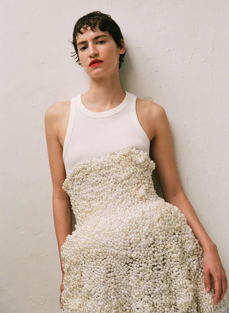 Fashionable young woman poses in a tank top and a dress entirely covered in white pearls against a white wall in a studio setting.