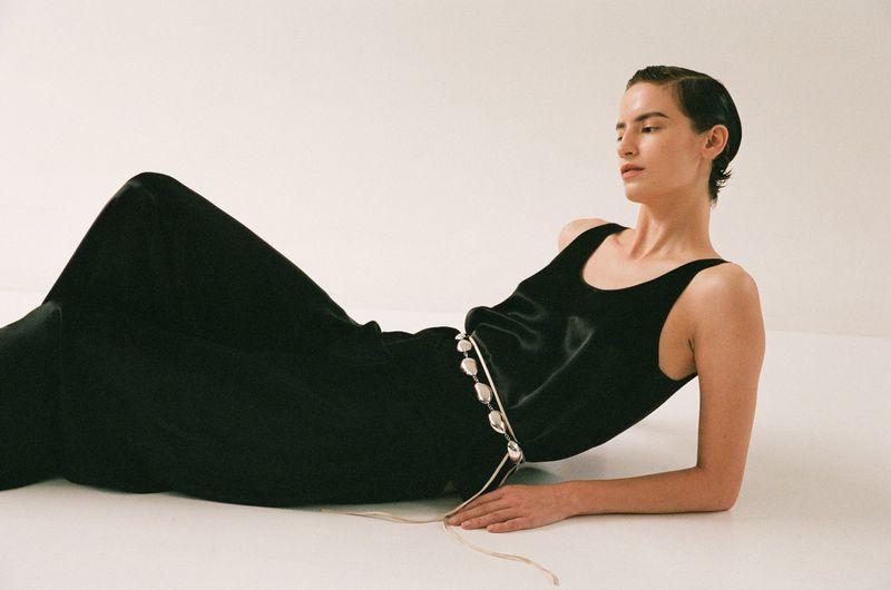 Fashion image of a slender adult female laying on a white surface, wearing a black top and long dress with a beaded belt.