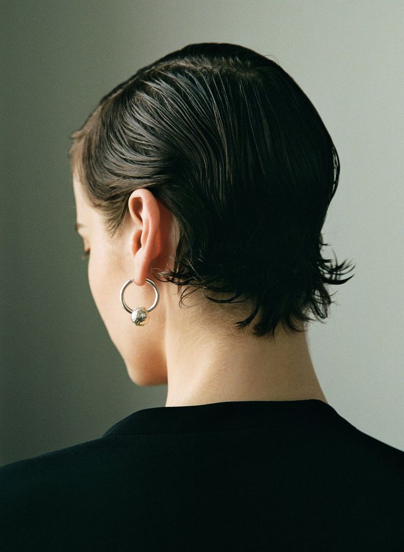 Back view of a female model with wet, dark, short hair wearing a silver hoop earring and black blazer against a gray background.