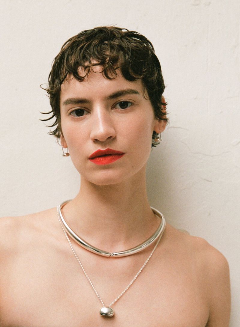 Close-up of a young adult female with short curly brown hair, wearing a silver collar necklace and red lipstick against a white wall.