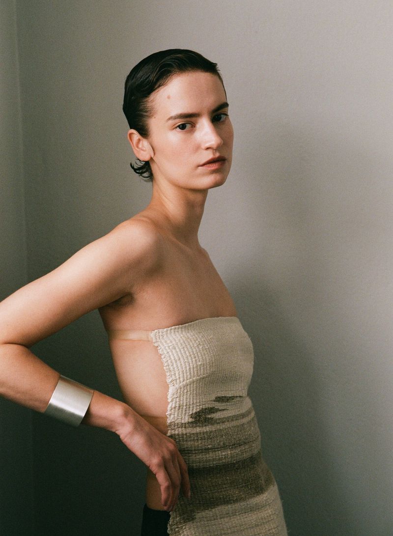 Portrait of a young woman wearing a tube top and silver cuff bracelet in front of a neutral wall, with short, slicked-back dark hair.