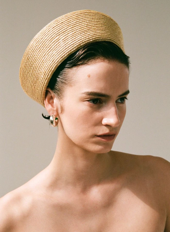 Model Stella Hägerstrand wearing a sculptural woven straw hat and gold hoop earring against a neutral backdrop.